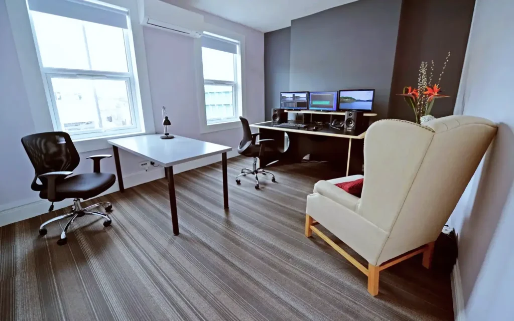Edit suite with three monitors, near-field speakers and an armchair, lit by two sash windows