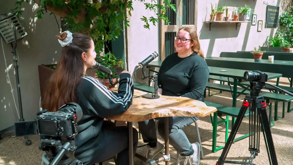 Two people recording a podcast outdoors at a courtyard table, with microphones and two cameras on tripods